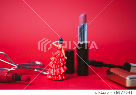 Decorative cosmetics products on red background - black mascara in gold tube, pink and red lipsticks, eyelash tongs. A toy woman in a red long dress with black hair. Female beauty and grooming concept 86977641