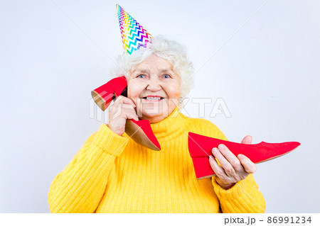 older woman with a gift wear yellow sweater and horn cap on a white background holding red high heels shoes 86991234