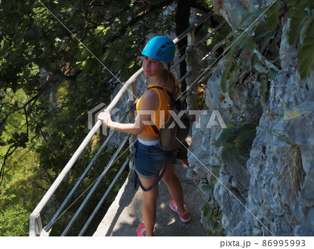 A girl in a helmet stands holding on to the railing on a narrow path of a rocky cliff in a green grove A girl in a helmet stands holding on to the railing on a narrow path of a rocky cliff in a green grove 86995993