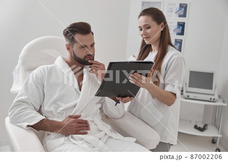 Administrator shows clipboard with form to client sitting in armchair in cosmetological clinic 87002206