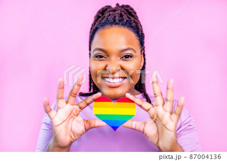 Portrait of an attractive hispanic woman holding a LGBT rainbow heart in pink studio 87004136
