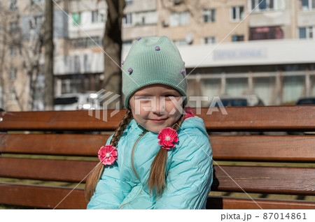 Portrait of a 5-year-old girl sitting on a park bench. Shy girl with long hair braided into pigtails. A child in a blue jacket and hat. Springtime. Portrait of a 5-year-old girl sitting on a park bench. Shy girl with long hair braided into pigtails. A child in a blue jacket and hat. Springtime. 87014861