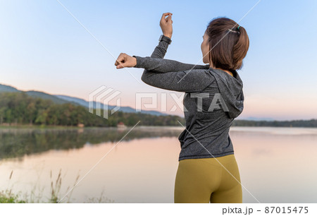Young attractive woman stretching her arms and legs before her morning exercise at a local lake park 87015475