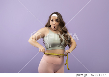 Surprised plus size european millennial woman in sportswear with open mouth measures waist with measuring tape 87021537