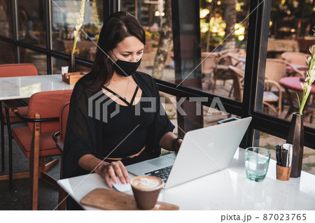 Young woman freelancer with protective medical black mask on face working with laptop in cafe Young woman freelancer with protective medical black mask on face working with laptop in cafe 87023765