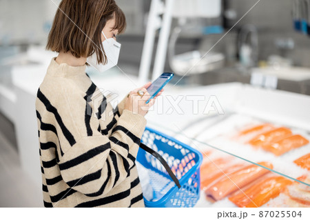 Woman in face mask using phone while standing with a shopping basket at shop 87025504