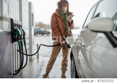 Woman in face mask refueling car with a gasoline using smart phone Woman in face mask refueling car with a gasoline using smart phone 87025522