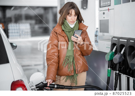 Woman in face mask refueling car with a gasoline using smart phone Woman in face mask refueling car with a gasoline using smart phone 87025523