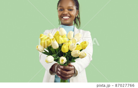 Happy Afro American girl holding a bunch of spring flowers and smiling. Closeup studio shot of a beautiful bouquet of fresh yellow and white tulips in the hands of a pretty young black woman 87027766