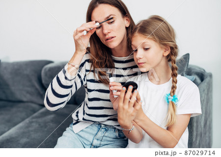 Mother applies mascara to her eyelashes looking into her daughters small mirror 87029714