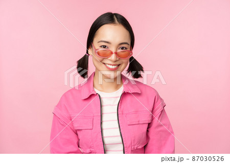 Close up of beautiful asian female model in stylish sunglasses, posing against pink background in trendy outfit, copy space 87030526
