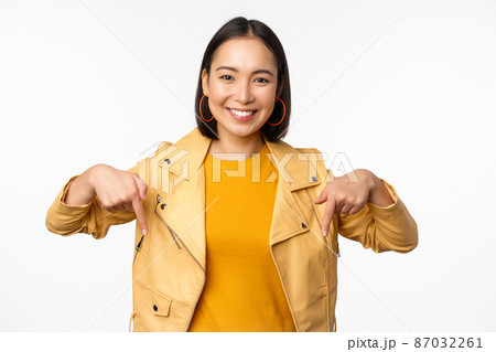 Portrait of happy smiling asian girl, pointing fingers down and showing logo, demonstrating banner, standing in yellow jacket against white background 87032261