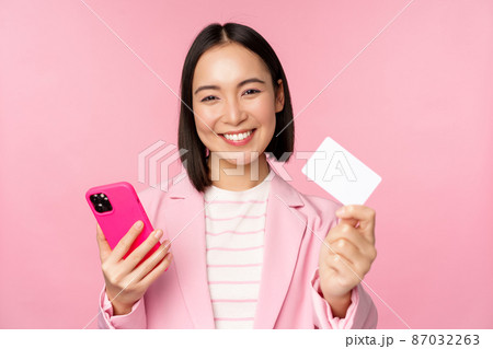 Image of smiling happy asian businesswoman showing credit card, paying online on smartphone application, order with mobile phon, standing against pink background 87032263