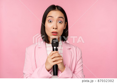Young asian saleswoman, office lady with suit, holding microphone and looking shocked at camera, talking, giving speech, standing over pink background 87032302