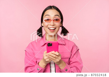 Portrait of korean girl in stylish sunglasses holding mobile phone, using smartphone app, standing over pink background 87032418