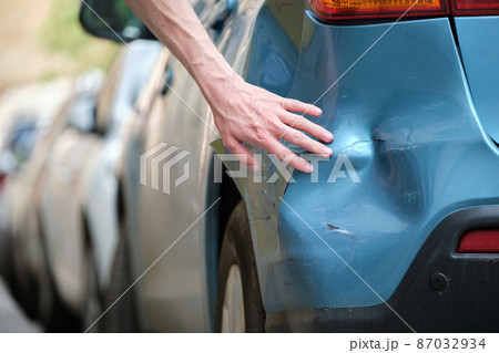 Driver hand examining dented car with damaged fender parked on city street side. Road safety and vehicle insurance concept Driver hand examining dented car with damaged fender parked on city street side. Road safety and vehicle insurance concept 87032934