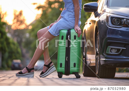 Closeup of young woman slim legs resting on suitcase bag beside car. Travelling and vacations concept. Closeup of young woman slim legs resting on suitcase bag beside car. Travelling and vacations concept. 87032980
