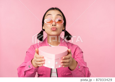 Cute asian girl giving you gift in heart shaped box, kissing and smiling, concept of holiday and celebration, standing over pink background 87033429