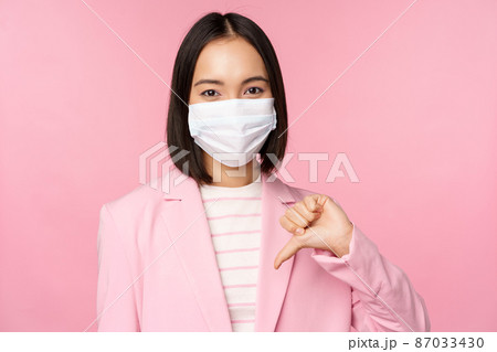 Korean business woman in medical face mask and suit, shows thumbs down, dislike or disapprove gesture, standing over pink background 87033430