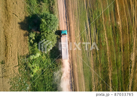 Aerial view of cargo truck driving on dirt road between agricultural wheat fields making lot of dust. Transportation of grain after being harvested by combine harvester during harvesting season Aerial view of cargo truck driving on dirt road between agricultural wheat fields making lot of dust. Transportation of grain after being harvested by combine harvester during harvesting season 87033765
