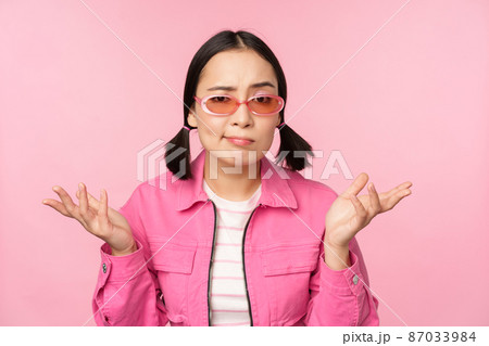 Close up portrait of asian girl looking confused, shrugging puzzled and looking at camera, wearing sunglasses, standing over pink background 87033984