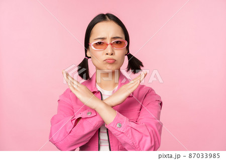 Portrait of korean girl in stylish sunglasses, sulking disappointed, showing stop, rejection gesture, cross sign, standing over pink background 87033985