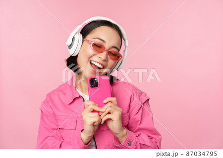 Stylish asian girl in headphones, listening music and taking photos on mobile phone, using smartphone, standing over pink background 87034795