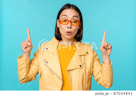 Enthusiastic asian girl in sunglasses, points fingers up, shows banner or logo on top, stands over blue background Enthusiastic asian girl in sunglasses, points fingers up, shows banner or logo on top, stands over blue background 87034841