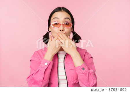 Portrait of shocked, stylish asian girl in sunglasses, closes mouth, looks with surprised face expression, stands over pink background 87034892