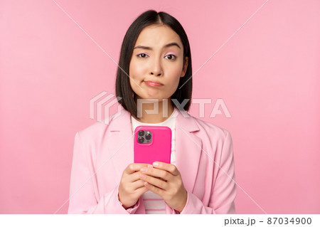 Doubtful businesswoman in suit, holding smartphone, grimacing, making skeptical face expression, standing over pink background 87034900