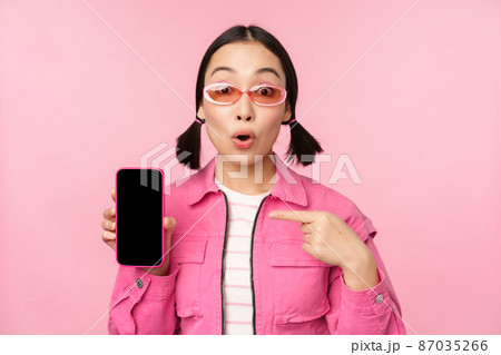Enthusiastic asian woman in stylish clothes, sunglasses, pointing finger at mobile phone screen, showing smartphone application, standing over pink background 87035266