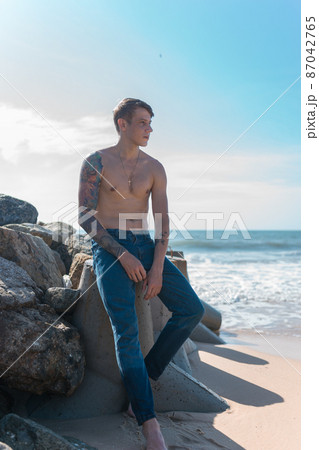 Handsome young caucasian bare-chested man with full-arm tattoo sitting on the rock by the sea. High quality photo Handsome young caucasian bare-chested man with full-arm tattoo sitting on the rock by the sea. High quality photo 87042765