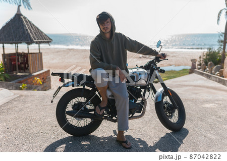 Young caucasian guy in hoodie sitting on motorcycle near the sea in a morning. Lovely sunshine. High quality photo Young caucasian guy in hoodie sitting on motorcycle near the sea in a morning. Lovely sunshine. High quality photo 87042822