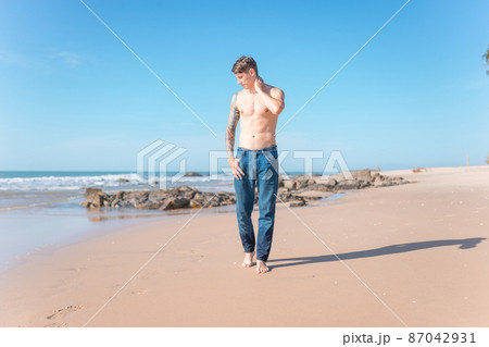 Attractive young caucasian bare-chested man with full-arm tattoo walking on the beach barefoot. Scenic ocean view, morning photo, wearing jeans. 87042931