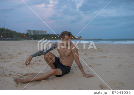 Full-length portrait of handsome caucasian man sitting on the sand on the beach on the sunset, full arm tattoo. High quality photo Full-length portrait of handsome caucasian man sitting on the sand on the beach on the sunset, full arm tattoo. High quality photo 87043059