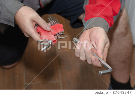 A man in workwear is holding a set of hex wrenches. An adult male with a hand tool in front of the brown wooden floor. A man in workwear is holding a set of hex wrenches. An adult male with a hand tool in front of the brown wooden floor. 87045247