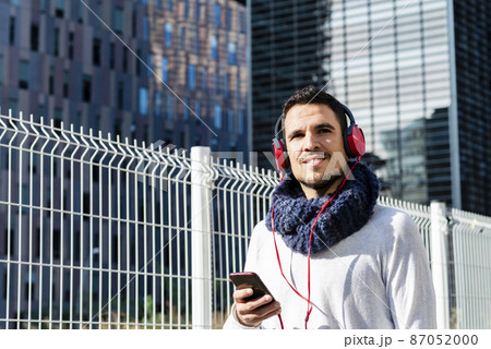 Young man listening music by headphones while holding a mobile phone outdoor 87052000