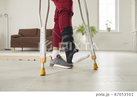 Injury and rehabilitation. Close up of legs of man who uses crutches to walk around room as he has leg injury. Uunknown man with ankle injury wears special bandage on his leg and uses metal crutches. Injury and rehabilitation. Close up of legs of man who uses crutches to walk around room as he has leg injury. Uunknown man with ankle injury wears special bandage on his leg and uses metal crutches. 87053501