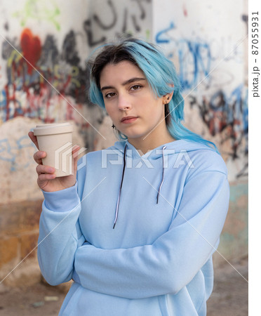 Blue haired Teenage girl in light blue oversize hoodie staying near graffiti wall with coffee to go 87055931