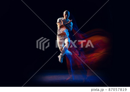 Jumping. Stylish man in sports white outfit dancing hip-hop, breakdance isolated on dark background in mixed neon light. Youth culture, hip-hop, style and fashion, action. 87057819