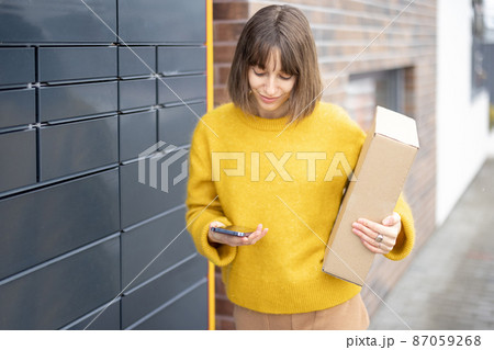 Happy woman with parcel and phone standing near automatic post terminal Happy woman with parcel and phone standing near automatic post terminal 87059268