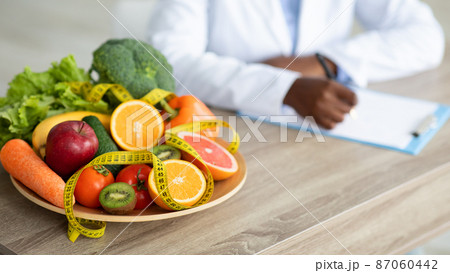 Black nutritionist writing diet plan, sitting at workplace in clinic, focus on plate with fruits and vegetables, cropped 87060442