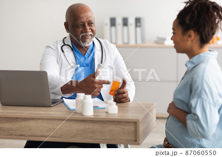 Mature experienced black doctor showing pills to pregnant lady 87060550