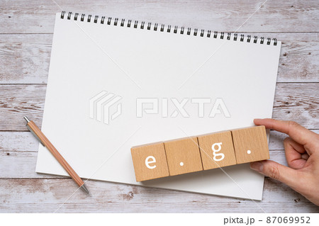 「e.g.」と書かれた積み木、ノート、ペン、人の手 87069952