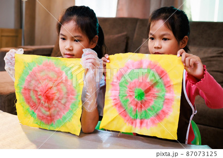 Asian Siblings child girl enjoy making tie dye cloth at home. Children show her finish tie dye cloth to her teacher and friend while online learning. Art and craft DIY for kid concept. 87073125