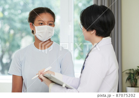 Asian doctor in white coat talking to girl in mask and filling the medical card during their conversation at hospital 87075202