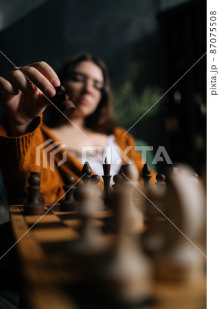 Vertical selective focus shot of confident businesswoman in elegant eyeglasses making chess move sitting on armchair in dark room. Pretty intelligent lady playing logical board game alone at home. Vertical selective focus shot of confident businesswoman in elegant eyeglasses making chess move sitting on armchair in dark room. Pretty intelligent lady playing logical board game alone at home. 87075508