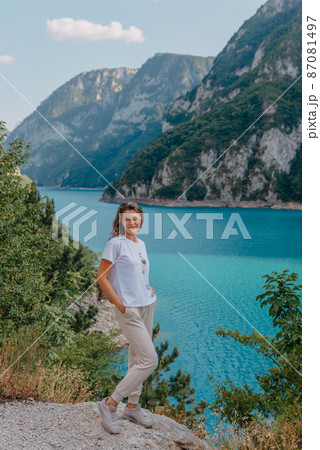 Girl tourist stands on the shore of a lake in the mountains. beautiful landscape, Piva Lake in Montenegro.. Woman is standing on the coast of Piva lake at sunset in summer. Landscape with girl, famous 87081497