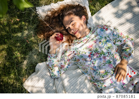 Portrait of beautiful woman posing lying on plaid at summer garden park. A book, fruit, a bag and a watermelon are next to each other. Portrait of beautiful woman posing lying on plaid at summer garden park. A book, fruit, a bag and a watermelon are next to each other. 87085181