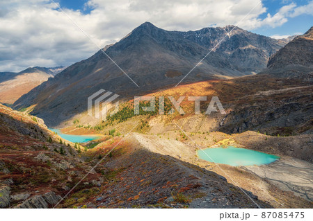 Dramatic foggy landscape with sunlit sharp mountain ridge with blue glacier lakes under low clouds at changeable weather. Atmospheric autumn mountain scenery with large sharp rocks on ridge top. Dramatic foggy landscape with sunlit sharp mountain ridge with blue glacier lakes under low clouds at changeable weather. Atmospheric autumn mountain scenery with large sharp rocks on ridge top. 87085475